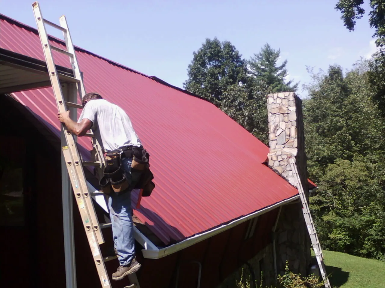 Roofer on a ladder beside a red metal roof and stone chimney — Soffit & Fascia Repair by Roofing Brothers in Falkland